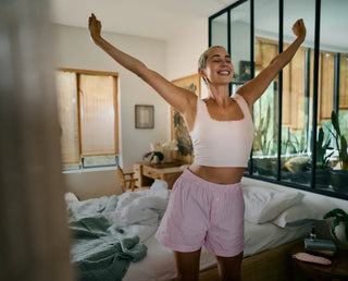 Eine Person in Tanktop und Shorts steht in einem sonnendurchfluteten Schlafzimmer neben einem ungemachten Bett, streckt die Arme über den Kopf und lächelt.
