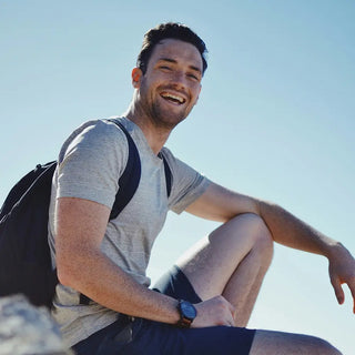 Un uomo in maglietta grigia, pantaloncini e zaino è seduto all'aperto su una roccia e sorride alla macchina fotografica sotto un cielo azzurro limpido.