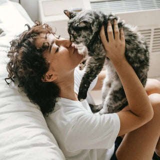 Persona seduta sul pavimento accanto a un letto in una stanza luminosa, che tiene un gatto grigio e nero al viso.