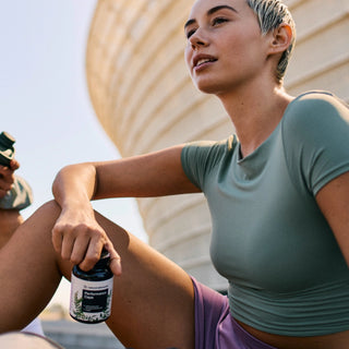 Una persona in abbigliamento sportivo è seduta all'aperto e tiene in mano un contenitore con la scritta "Performance Capsules"; sullo sfondo è visibile un moderno edificio curvo.