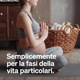 Una donna incinta in abbigliamento sportivo è seduta a gambe incrociate su un tappetino da yoga, con le mani in posizione di preghiera; cesto di vimini e asciugamani sullo sfondo. Testo italiano sovrapposto all'immagine.
