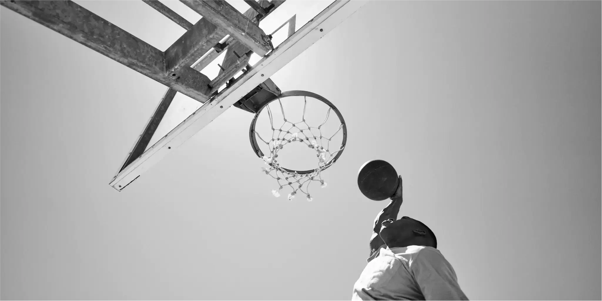 Eine Person, die gerade dabei ist, einen Basketball in einen Korb im Freien zu werfen, von unten vor einem klaren Himmel gesehen.