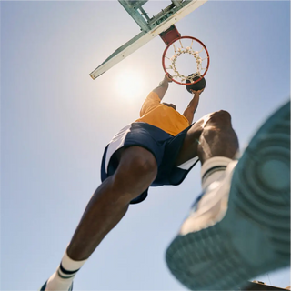 Untersicht eines Basketballspielers, der in der Luft einen Ball durch einen Korb dunkt, während hinter ihm die Sonne scheint.