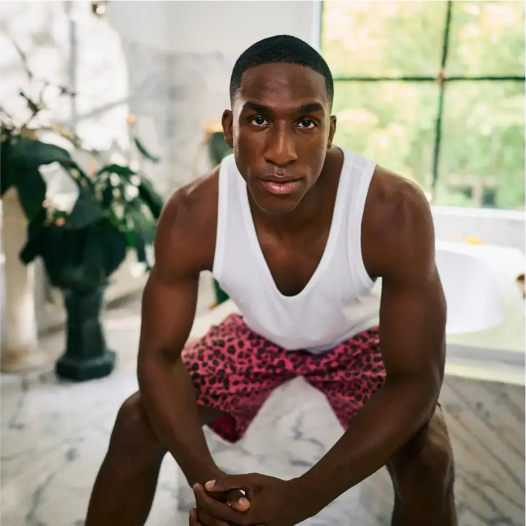 Ein Mann in einem weißen Tanktop und rosa Shorts mit Leopardenmuster sitzt mit gefalteten Händen auf einem Marmorboden im Innenbereich. Im Hintergrund sind große Fenster und Pflanzen zu sehen.