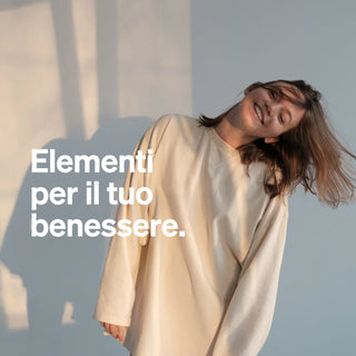 Una donna sorridente con un maglione color crema si trova davanti a una parete chiara, su cui è visualizzato il testo in italiano "Elementi per il tuo benessere".