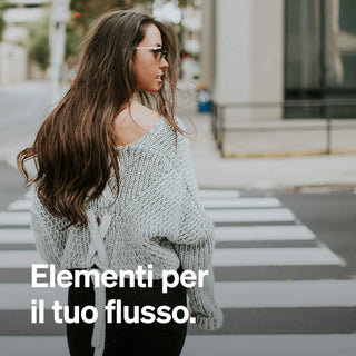 Eine Frau mit langen braunen Haaren und einem lockeren Pullover überquert eine Straße in der Stadt. Der weiße Text auf Italienisch lautet: "Elementi per il tuo flusso.