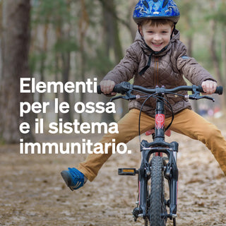 Un bambino piccolo con il casco va in bicicletta con le gambe tese su un sentiero nel bosco. Il testo in italiano promuove la salute delle ossa e del sistema immunitario.
