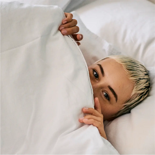 Person mit kurzen blonden Haaren liegt im Bett, ist teilweise bis zur Nase mit einer weißen Decke bedeckt und blickt direkt in die Kamera.