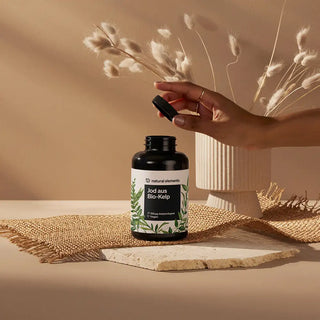 Eine Hand öffnet eine Flasche mit der Aufschrift "Jod aus Bio-Kelp" von Natural Elements, die auf einer strukturierten Matte mit getrockneten Pflanzen im Hintergrund steht.