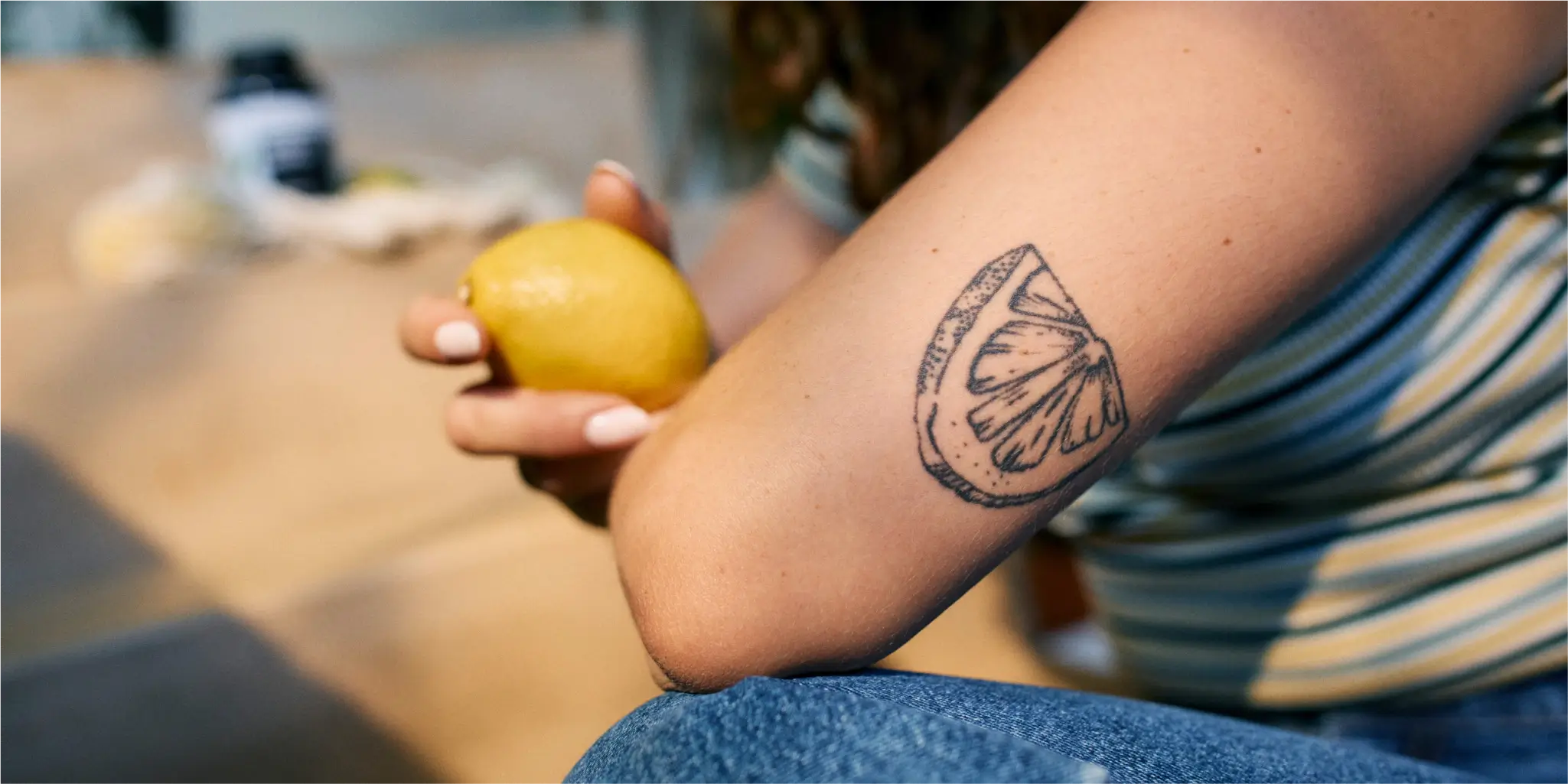 Person, die eine Zitrone hält und auf dem Unterarm eine Tätowierung einer Zitronenscheibe hat; sie trägt ein gestreiftes Hemd und Jeans.