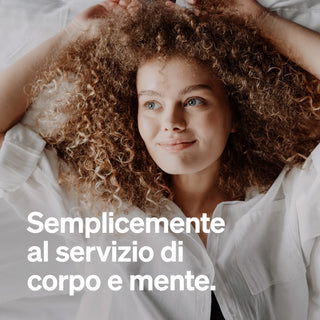 Una donna con i capelli ricci è sdraiata su un letto, guarda in alto e sorride leggermente. Il testo italiano recita: "Semplicemente al servizio di corpo e mente."