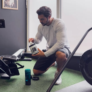Un uomo è inginocchiato sul pavimento di una palestra e preleva della polvere da un contenitore versandola in uno shaker, accanto a un vogatore.