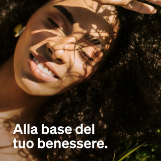 Una donna giace al sole con gli occhi chiusi e sorride, mentre ombre di foglie le cadono sul viso. Il testo recita: "Alla base del tuo benessere."