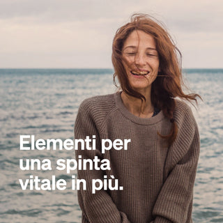 Una donna con lunghi capelli rossi sorride in una giornata nuvolosa al mare. Indossa un maglione marrone. Il testo italiano recita: "Elementi per una spinta vitale in più."