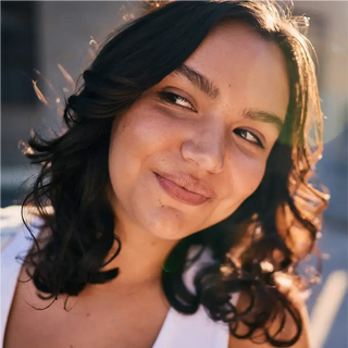 Una persona con capelli mossi e scuri e un top bianco sorride leggermente mentre guarda di lato alla luce del sole.