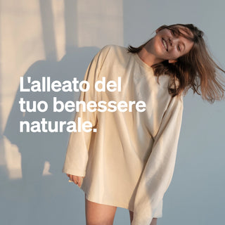 Una donna sorridente in una camicia beige oversize è in piedi in un ambiente interno, dove la luce del sole proietta ombre sul muro. Il testo italiano recita: "L'alleato del tuo benessere naturale."