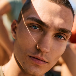 Nahaufnahme einer jungen Person mit kurzen Haaren und blauen Augen, die direkt in die Kamera blickt, wobei das Sonnenlicht einen Schatten auf ihr Gesicht wirft.