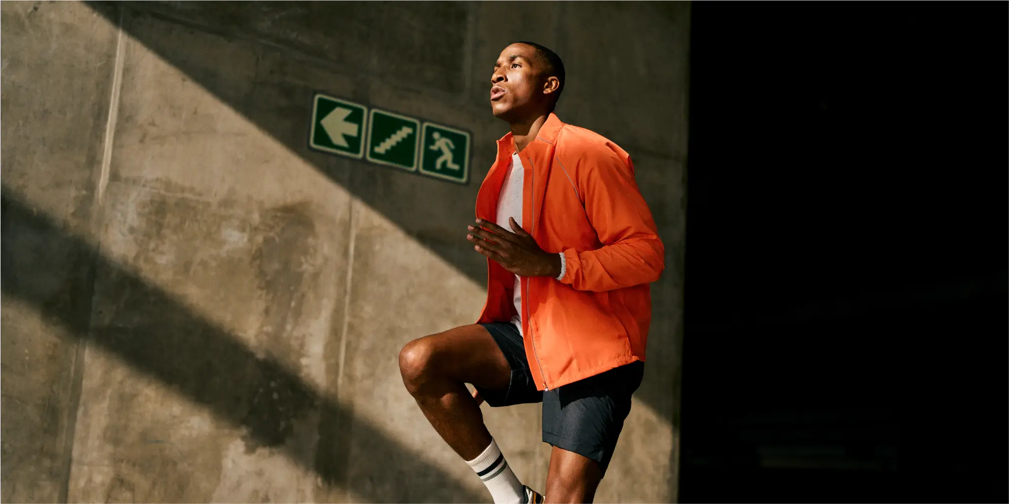 Ein Mann in Sportkleidung joggt in einem Gebäude neben einer Betonwand mit Notausgangs- und Treppenschildern, die vom Sonnenlicht beleuchtet wird.