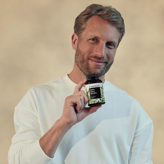 Ein Mann im Sweatshirt hält eine Flasche des Nahrungsergänzungsmittels "Omega Complex Lemon" mit natürlichen Bestandteilen in die Höhe und lächelt in die Kamera.