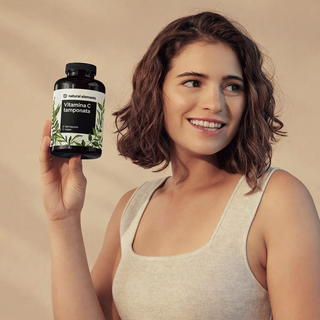 Eine Frau in einem beigefarbenen Tanktop hält lächelnd eine Flasche "Vitamin C tamponata" von Natural Elements hoch.