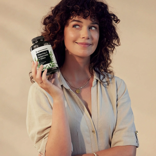 Eine Frau mit lockigem Haar hält eine Flasche mit Vitamin C, Kurkuma und Zinkpräparaten in der Hand, lächelt und blickt zur Seite.