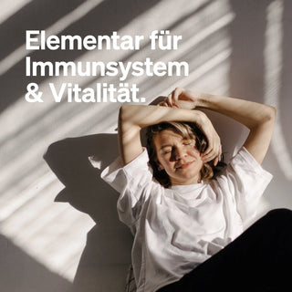 Frau in weißem Shirt liegt entspannt am Boden, daneben der deutsche Text: "Elementar für Immunsystem & Vitalität".
