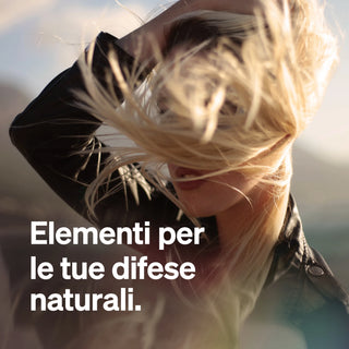 Una persona con lunghi capelli biondi, che le coprono parzialmente il viso, sta all'aperto alla luce del sole e indossa una giacca nera. Il testo italiano recita: "Elementi per le tue difese naturali."