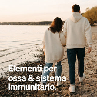 Due persone in felpa con cappuccio camminano lungo una spiaggia rocciosa al tramonto. Il testo in italiano recita: "Elementi per ossa & sistema immunitario."