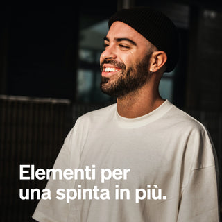 Lächelnder Mann mit Bart in weißem T-Shirt und schwarzer Mütze. Italienischer Text: "Elementi per una spinta in più.