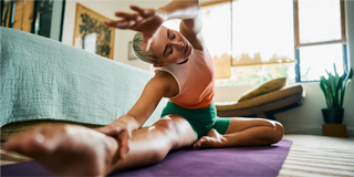 Persona seduta su un tappetino da yoga in un soggiorno, che allunga un braccio sopra la testa e si protende verso la gamba estesa.