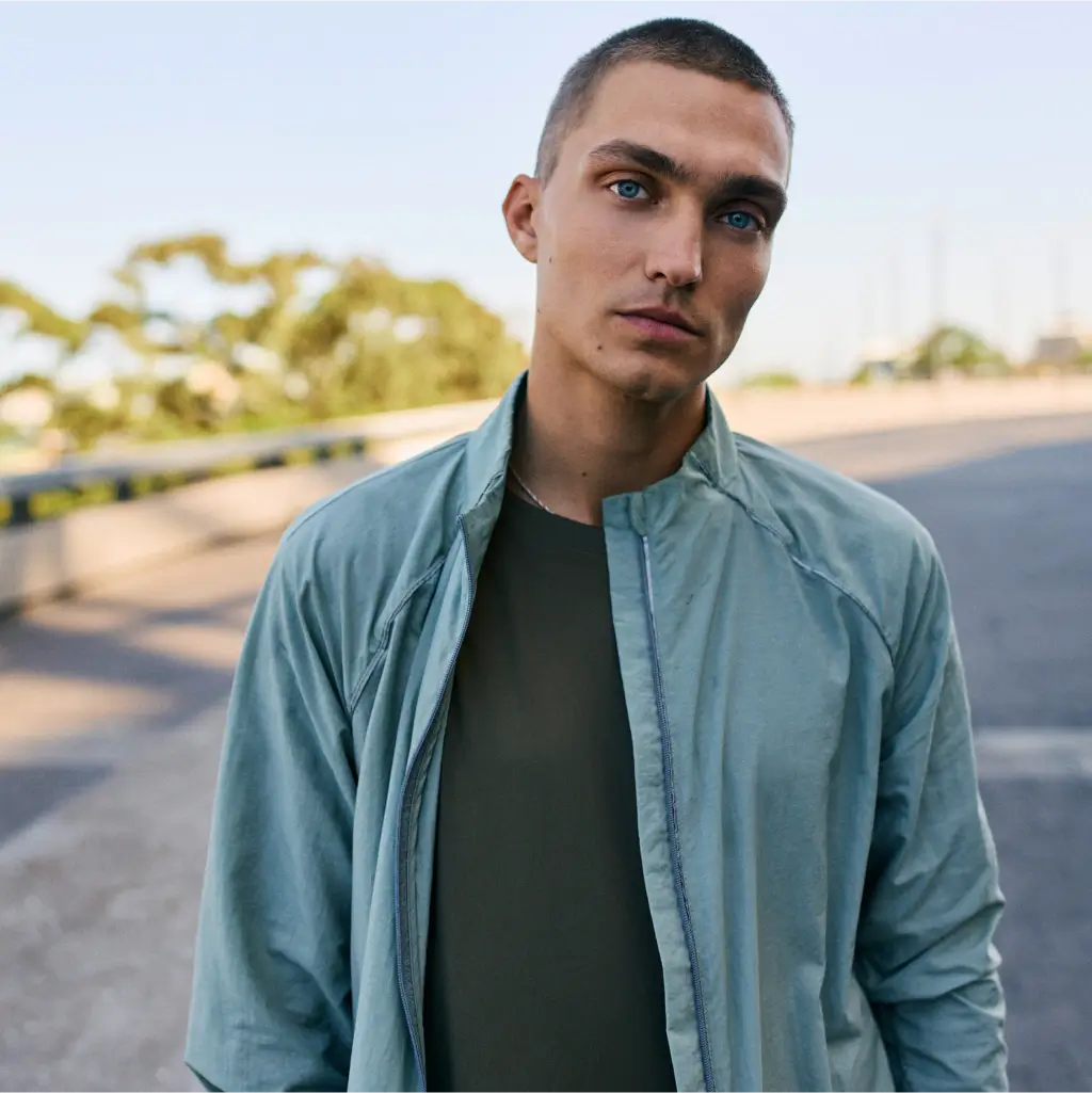 Ein junger Mann mit kurzen Haaren, der eine hellblaue Jacke über einem dunklen T-Shirt trägt, steht im Freien auf einer gepflasterten Fläche mit verschwommenen Bäumen im Hintergrund.
