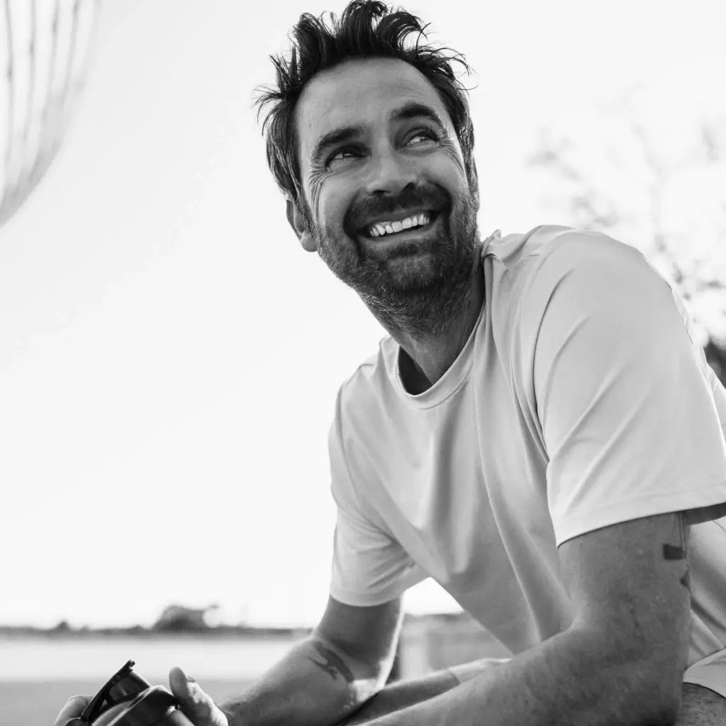 Ein Mann mit Bart und zerzaustem Haar sitzt lächelnd draußen und hält eine Kamera in der Hand. Er trägt ein helles T-Shirt. Das Bild ist schwarz-weiß.
