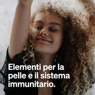 Una persona con i capelli ricci sorride con gli occhi chiusi e le braccia alzate. Il testo italiano sopra l'immagine recita: "Elementi per la pelle e il sistema immunitario."