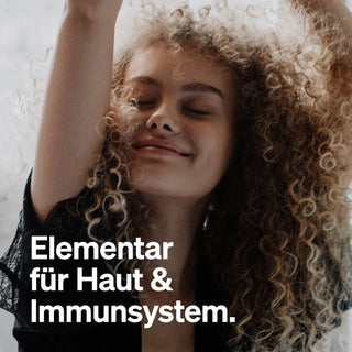 Eine Person mit lockigem Haar lächelt mit geschlossenen Augen und strahlt Freude aus. Der Text: "Elementar für Haut & Immunsystem" hebt natürliche Elemente hervor.